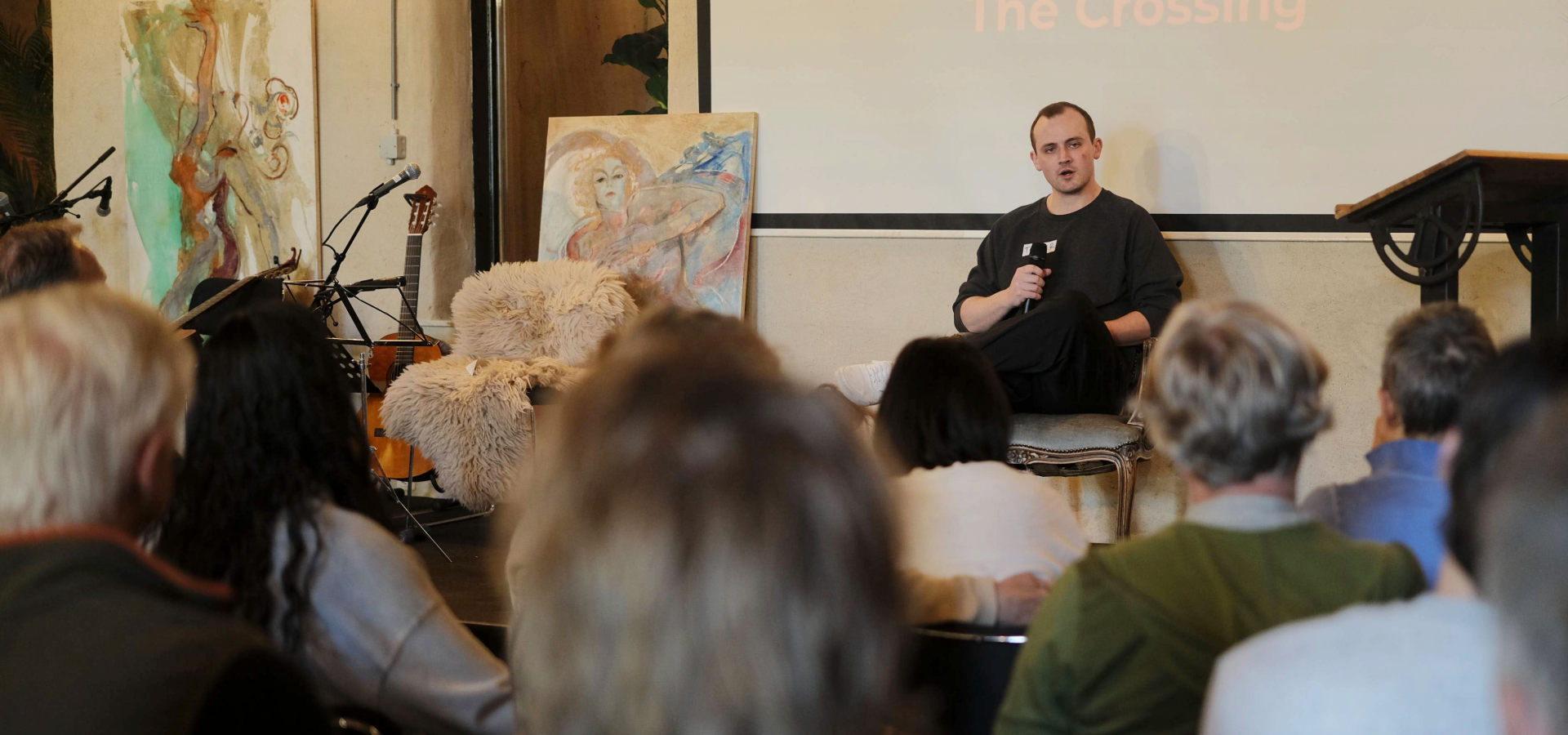 A teacher seated with a microphone before a full room, The Crossing shown on the screen behind