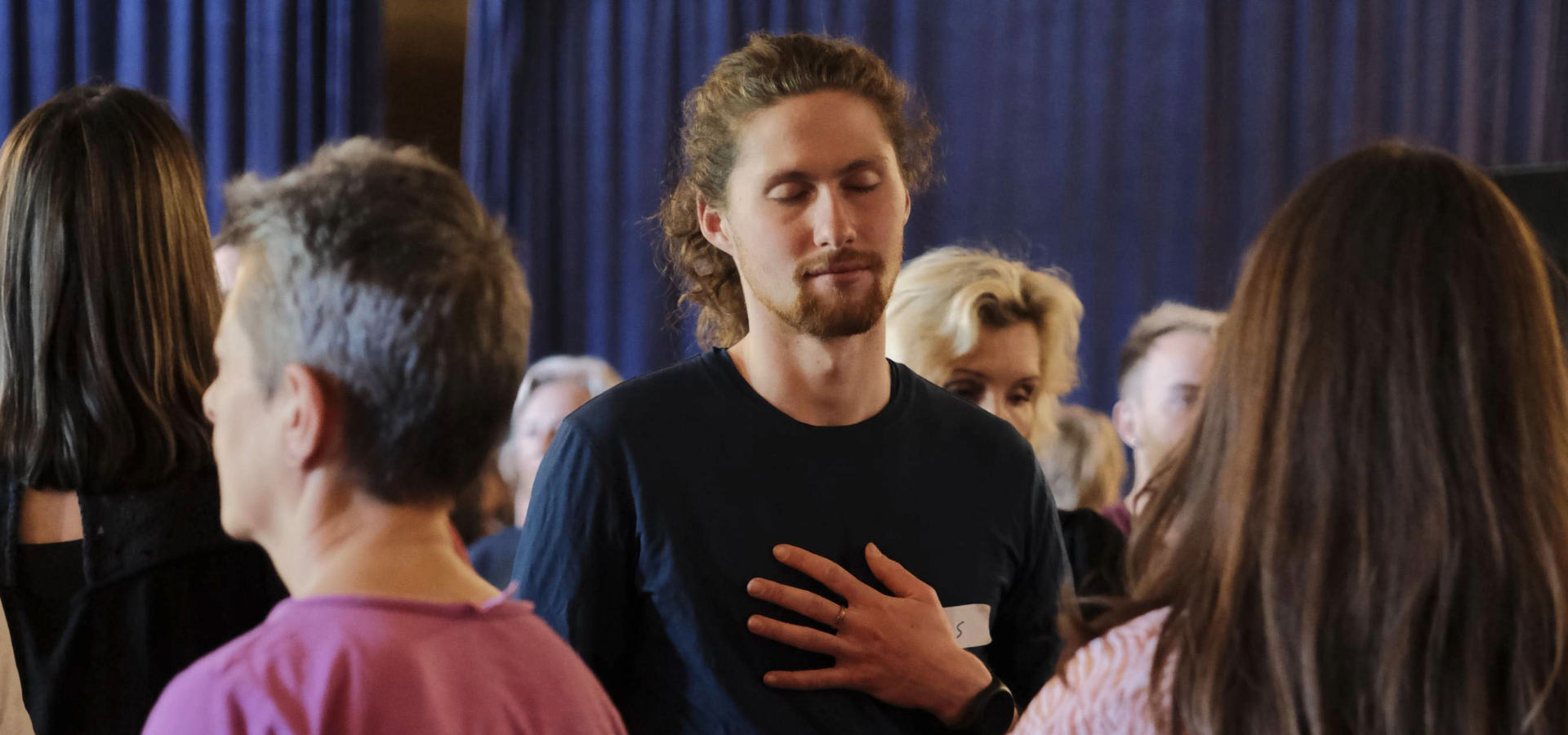 A young man standing with his eyes closed and his hand on his chest, held within a circle of people